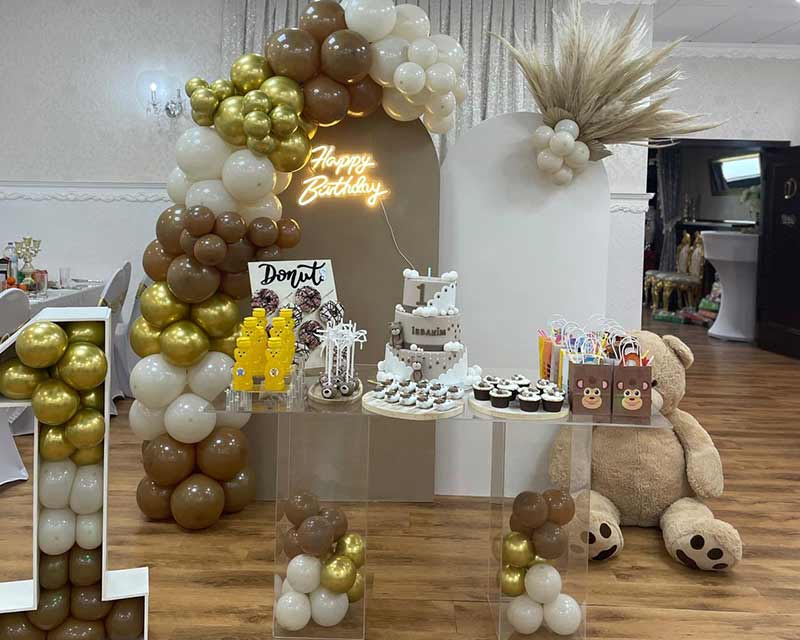 Geburtstagstisch mit goldenen und weißen Ballons, Donuts und einer Torte.